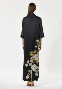 Kvinde iført en lang sort kjole med beige blomsterprint på den nederste del af ryggen og guld højhælede sko, stående med ryggen til.