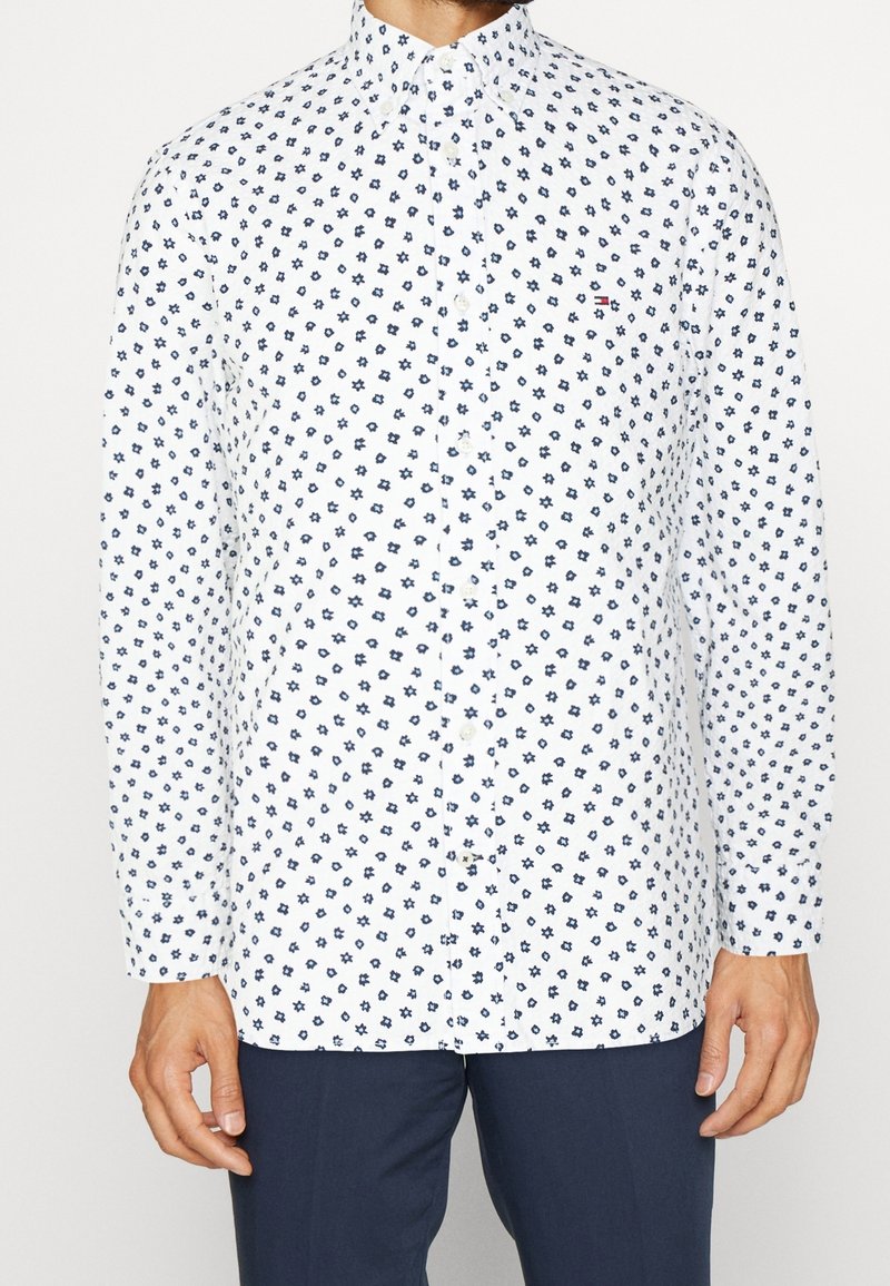 Homme portant une chemise blanche à boutons avec de petits motifs floraux bleu marine et un pantalon bleu foncé, debout les bras relâchés.
