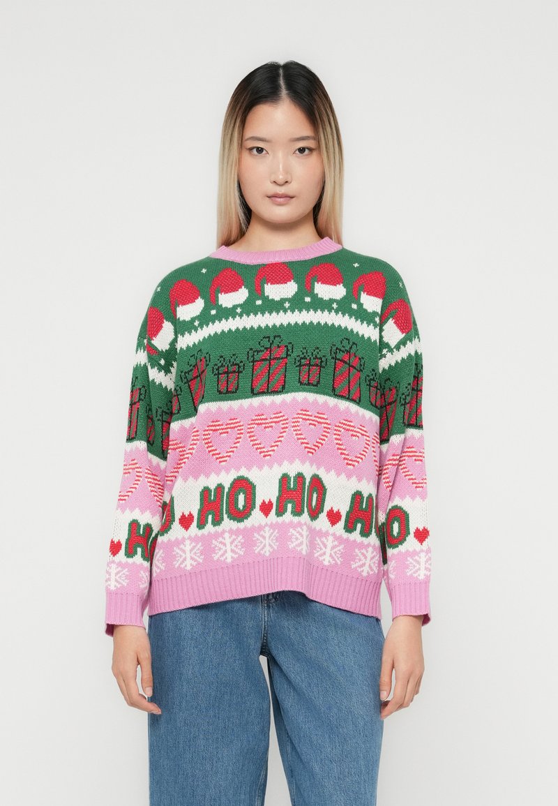 Unge kvinde iført en farverig ju sweatshirt med julemandshatte, gaveæsker, hjerter, snefnug og teksten "HO HO HO", der er parret med blå jeans.