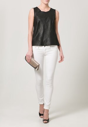 Mujer que lleva una blusa de cuero negra sin mangas, jeans ajustados blancos, tacones negros abiertos en los dedos, sosteniendo un bolso de mano beige.