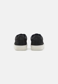 Black suede sneakers with white rubber soles, featuring a smooth texture and minimalistic design. Rear view highlights rounded shoe back.