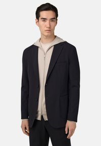 Námořnický blazer s dvěma knoflíky a kapsami, vrstvený přes světle béžovou mikinu na zip. Hladká tkanina, přizpůsobený střih a moderní design.