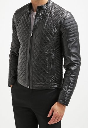 Chaqueta de cuero negra acolchada con cierre de cremallera frontal y bolsillos con cremallera, usada por una persona con las manos en los bolsillos, combinada con pantalones negros.