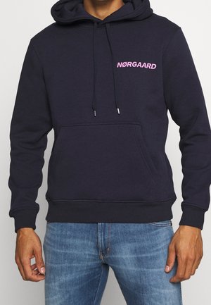 Homme portant un sweat à capuche bleu marine avec une poche devant et un texte rose "NØRGAARD" sur la poitrine gauche, associé à un jean bleu.