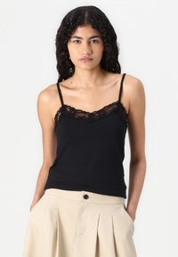 Svart cami med blondekant rundt halsen, med tynne stropper og en tettsittende design, kombinert med et lys beige plisserte skjørt.