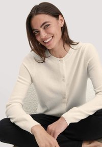 Cremfarbener Cardigan aus weichem Material mit rundem Halsausschnitt, fünf Knöpfen auf der Vorderseite und gerippten Bündchen. Getragen mit schwarzen Hosen.
