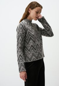 Grauer gestrickter Pullover mit einem Zickzackmuster in Schwarz und Grau, mit hohem Kragen und langen Ärmeln. Der Stoff wirkt weich und strukturiert.