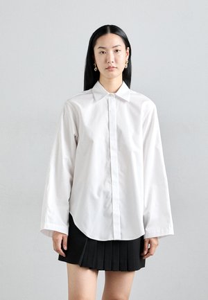 White long-sleeve shirt in a crisp material with a button-down front, flat collar, and a rounded hem, paired with a black pleated skirt.