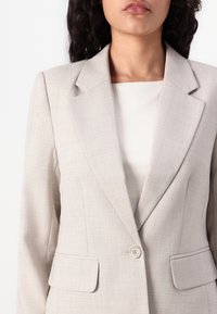 Blazer bej deschis dintr-un material texturat. Are rever notat, închidere cu un singur nasture și două buzunare frontale. Captuseala interioara este netedă.