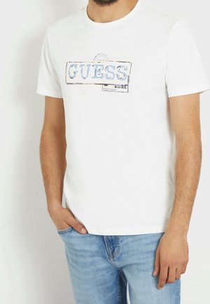 T-shirt en coton blanc avec un logo "GUESS" bleu et orange encadré dans un rectangle, manches courtes et col rond.