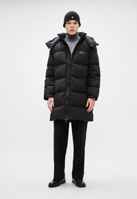 Black puffer coat with a zip front, large hood, and a logo patch. Worn over a gray turtleneck and black trousers. Smooth texture.