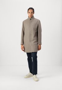 Brun ullfrakk med høy krage, frontlukke med knapper og to sidelommer, kombinert med mørke jeans og hvite sneakers.