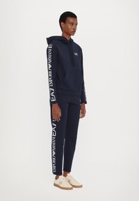 Sudadera con capucha y joggers en color navy, con detalles blancos en las mangas y piernas, hechos de una tela suave. Zapatillas beige con suelas marrón claro.