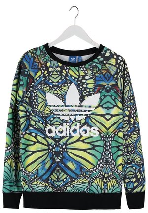 Osoba nosząca kolorową bluzę Adidas, ciemne obcisłe jeansy, białe wysokie trampki Adidas i trzymająca niebieski plecak Adidas za uchwyt.