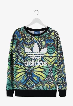 Persoon draagt een kleurrijk Adidas sweatshirt, donkere skinny jeans, witte Adidas hoge sneakers en houdt een blauwe Adidas rugzak aan het handvat vast.