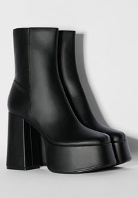 Un par de botines de plataforma de cuero negro con tacones altos gruesos, punteras redondeadas y un diseño sin costuras sobre un fondo claro.