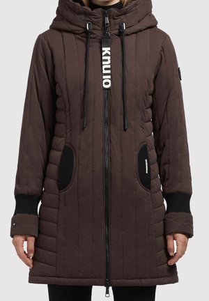 Femme portant un manteau d'hiver matelassé marron avec capuche, poignets élastiques noirs, fermeture éclair avant et poches latérales noires avec des patchs texturés.