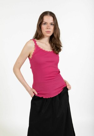 Mlada ženska z dolgimi rjavimi lasmi, oblečena v roza zgornji del brez rokavov in črno krilo, stoji z rokami v žepih na belem ozadju.