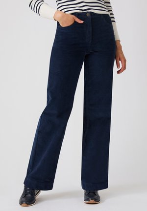 Vrouw die marineblauwe wijde broek, zwarte sneakers en een witte trui met zwarte strepen draagt, staat met één hand in haar zak.