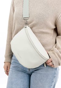 Weiße Leder-Crossbody-Tasche mit glatter Textur, geschwungenem Design und grauem verstellbarem Riemen. Verfügt über einen Reißverschluss und minimalistisches Branding.