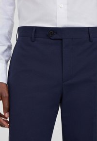 Pantaloni blu navy realizzati in tessuto testurizzato, con chiusura frontale a bottone e passanti per la cintura, dalla vestibilità aderente e dal design slanciato.