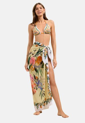 Kvinde iført en bikinitop med blomsterprint og en matchende sarong med frugt- og blomsterdesign bundet om livet, stående barfodet.