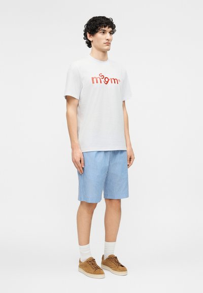 Νεαρός άντρας που φοράει λευκό μπλουζάκι με κόκκινο λογότυπο, μπλε και λευκές ριγέ βερμούδες, λευκές κάλτσες και καφέ αθλητικά παπούτσια, στέκεται μπροστά σε απλό φόντο.