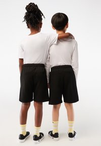 Deux enfants portant des hauts blancs, des shorts noirs, des chaussettes jaunes rayées et des chaussures noires, se tiennent côte à côte, un bras autour de l'épaule de l'autre, tournés de dos.