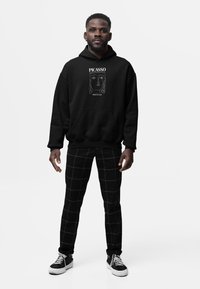 Černá mikina s kapucí a bílým potiskem, který zobrazuje Picassovu "Hlava dívky", v kombinaci s černými kostkovanými kalhotami a černými teniskami.