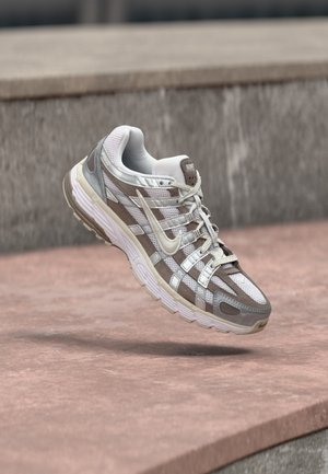 Hvid og grå Nike løbesko svævende over en lyserød betonoverflade med en grå stenmur i baggrunden.