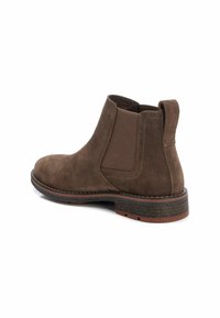 Botín de ante marrón con paneles elásticos en los laterales, puntera redondeada y suela de goma texturizada. Presenta detalles de costura y una lengüeta para facilitar el calce.