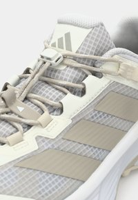Světle šedá sportovní obuv s textilním svrškem z mřížky, strukturovanými panely a třemi béžovými pruhy. Disponuje systémem na uchycení tkaniček a polstrovaným límcem.