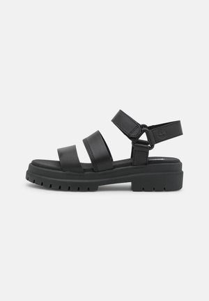 Sandale en cuir noir avec trois brides, bride de cheville réglable avec Velcro, et semelle épaisse à crampons pour une meilleure adhérence et un grand confort.