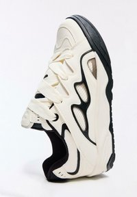 Fehér bőr felsőrész fekete díszítésekkel, hálós panelekkel és vastag talppal ellátott lábbeli. Láthatóak a fűzők és a párnázott nyakrész.