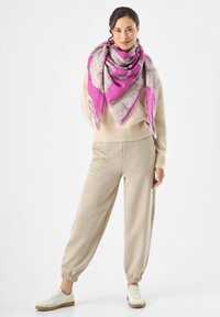 Sciarpa triangolare rosa e beige con un motivo texturizzato, indossata sopra un maglione beige e pantaloni slouchy abbinati, completata da sneakers bianche.