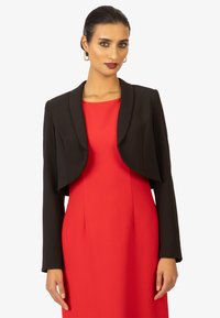 Blazer nero corto con colletto arrotondato, dotato di maniche lunghe. Indossato sopra un vestito rosso, mette in risalto un design strutturato e una trama morbida.