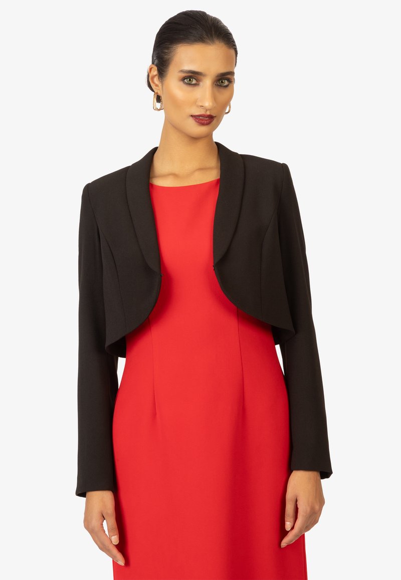 Blazer nero corto con colletto arrotondato, dotato di maniche lunghe. Indossato sopra un vestito rosso, mette in risalto un design strutturato e una trama morbida.