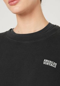 Black sweatshirt with a ribbed crew neck. Features a small white "AMERICAN VINTAGE" logo on the left chest area. Soft fabric texture.