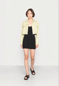 Vestido preto com textura em cordas, sobreposto por uma jaqueta cropped verde-clara. Sandálias abertas, tipo slip-on, pretas finalizam o visual. Design simples e moderno.