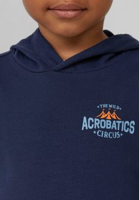 Navyblauer Hoodie aus Baumwolle mit einem frontalen Grafikdesign, das "The Wild Acrobatics Circus" in blauem und orangefarbenem Text sowie Zeltmotiven zeigt.
