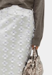 Falda blanca con apliques florales que presentan delicadas flores de tela y centros de strass, combinada con un suéter gris y un bolso texturizado con estampado de serpiente.