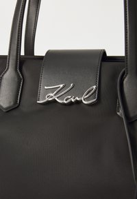 Black tote bag with smooth synthetic material, featuring a structured design, flat top, dual handles, and a silver 'Karl' accent on the flap.