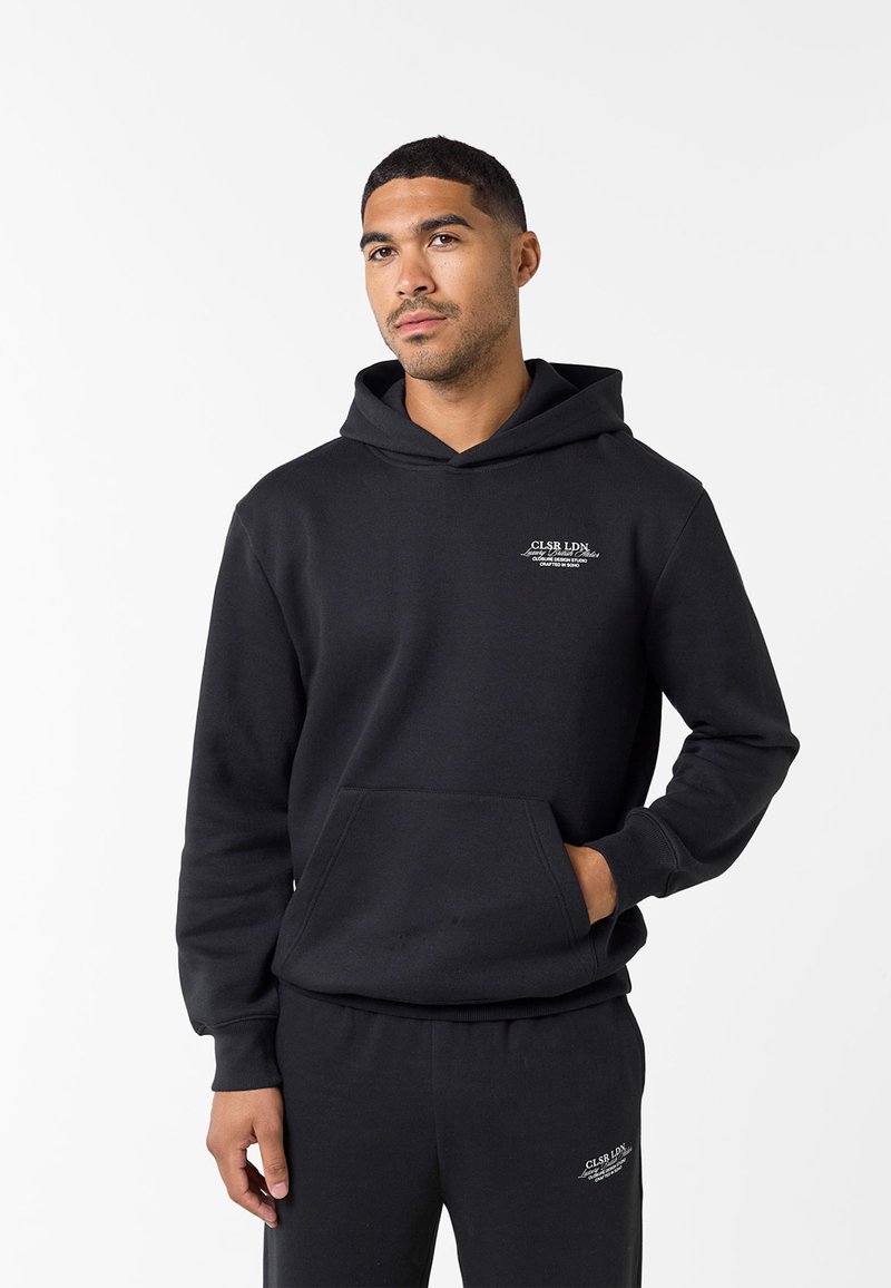 Άνδρας που φορά μαύρη φανέλα με κουκούλα και ταιριαστό παντελόνι φόρμας με μικρό λευκό κείμενο τυπωμένο στο στήθος και στο μηρό, στέκεται μπροστά σε λευκό φόντο.