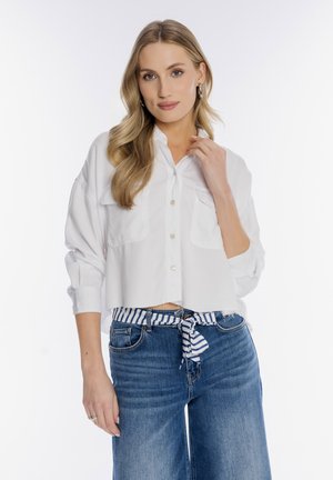 Blondynka z luźnymi falami ubrana w oversize'ową białą koszulę na guziki, niebieskie dżinsy i paskiem w niebiesko-białe paski, pozująca na białym tle.