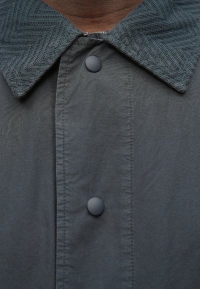 Szara kurtka zapinana na guziki z teksturowanym kołnierzem w jodełkę; posiada dwa czarne plastikowe guziki oraz gładkie wykończenie materiału.