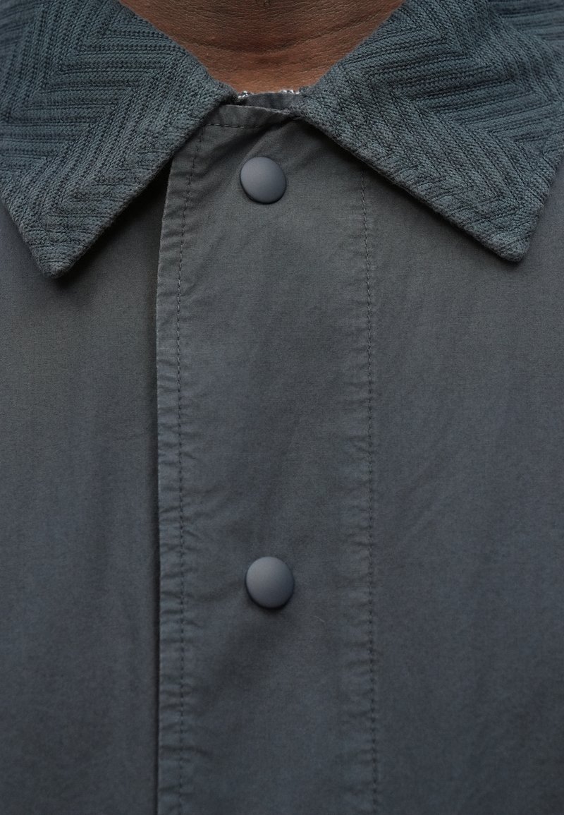 Gray button-up jacket with a textured, herringbone-patterned collar; features two black plastic buttons and a smooth fabric finish.