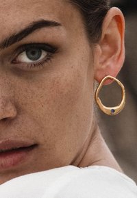 Auksiniai auskarai žiedai su organine forma, šiurkščiu paviršiumi ir mažu mėlynu akcentu. Užsidėta ant modelio ausies.