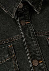 Chaqueta de mezclilla negra con dos bolsillos en el pecho, botones de color cobre y costuras naranjas que contrastan a lo largo de las costuras y el cuello.