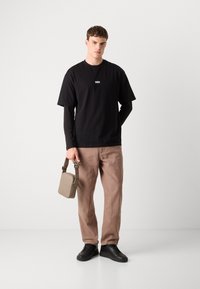 T-shirt nera con un piccolo logo bianco, indossata sopra una maglietta a maniche lunghe. Pantaloni beige e una piccola borsa a tracolla beige. Sneakers nere.