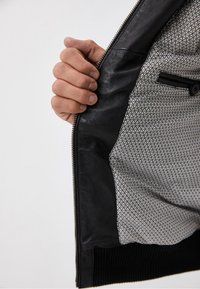 Interior de una chaqueta de cuero negra con un forro de tela texturizada que presenta un patrón geométrico. Cierre plateado y mano sosteniendo la abertura.